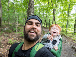 Photo: smiling man hiking in green woods with laughing baby in front carrier
