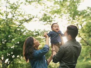 Photograph: parents lift smiling baby high outdoors under sunlit green trees