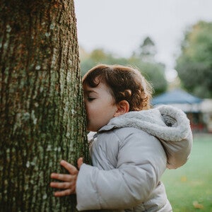 Photograph of a child in a gray coat pressing their face against a tree in a park
