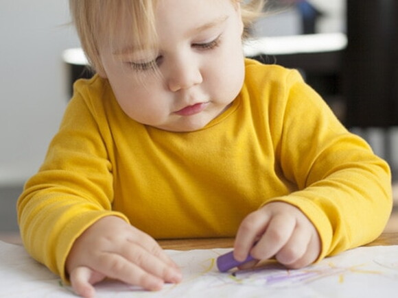 Bébé avec un pull jaune est assis à son bureau dans sa classe et est concentré à dessiner avec un crayola sur une feuille
