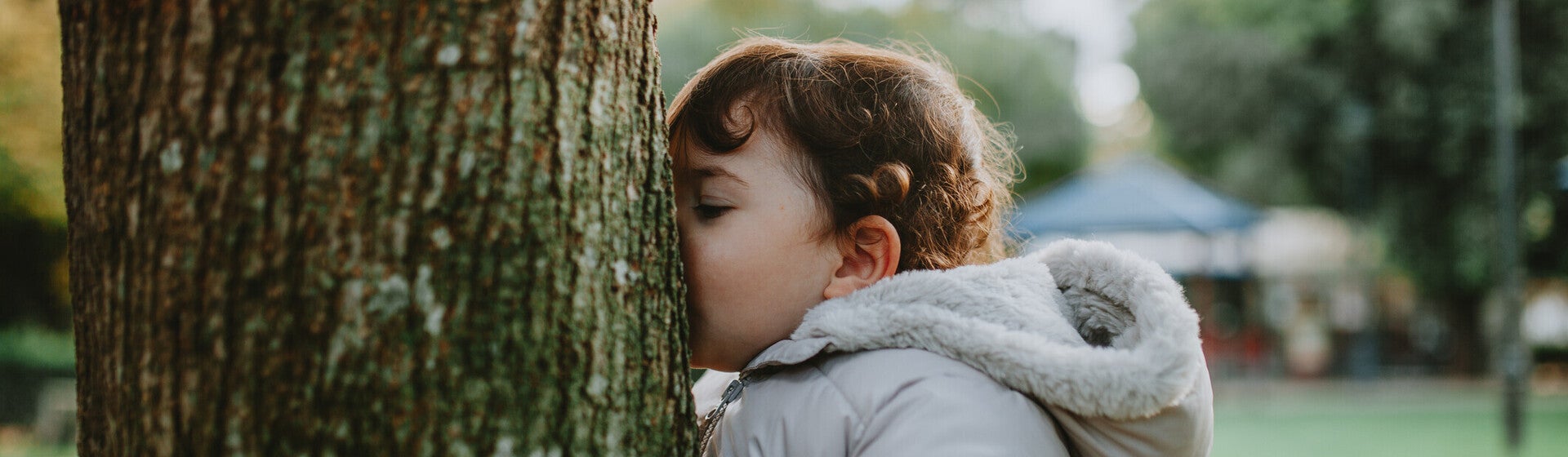 Photo of toddler in gray coat pressing cheek to tree in a green park